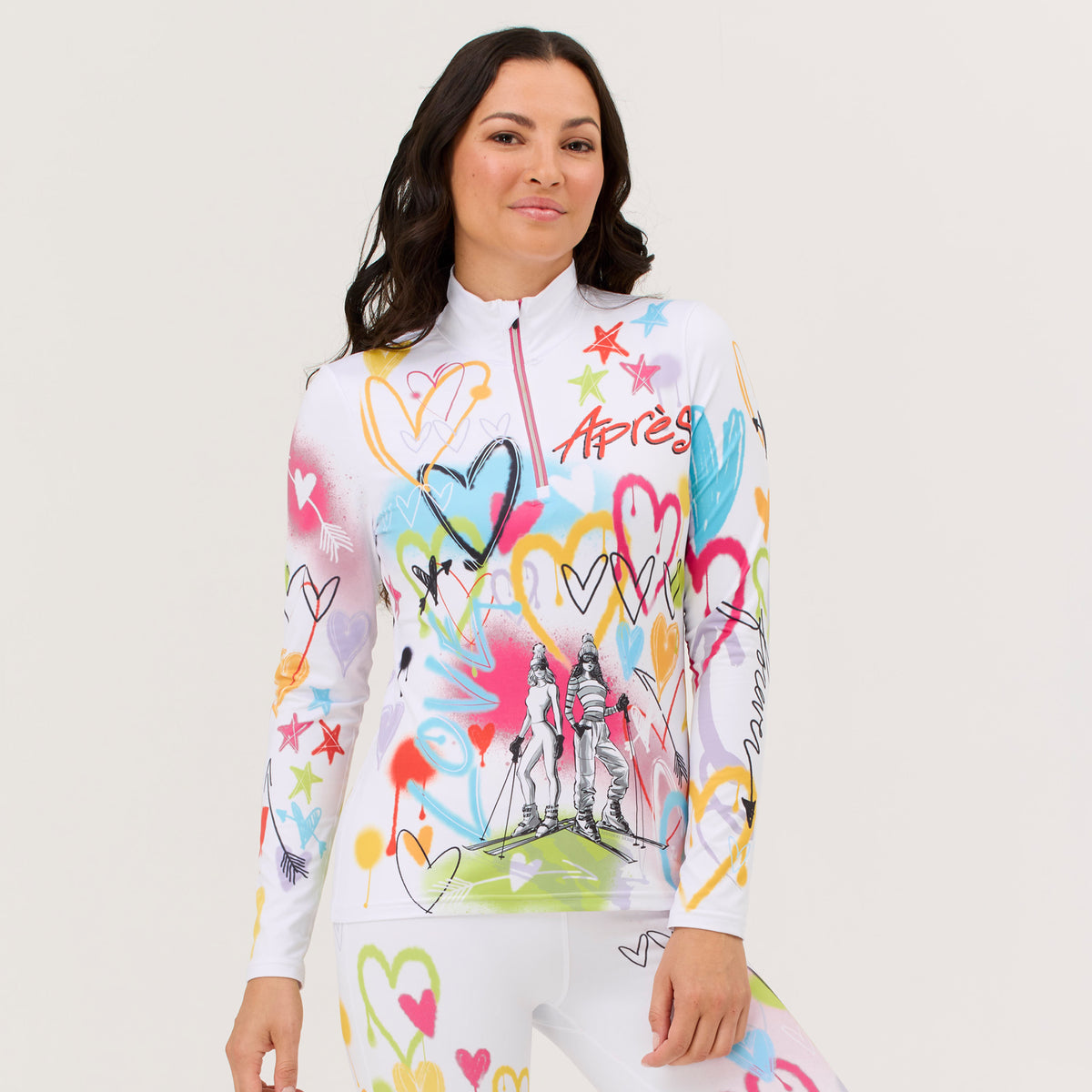 A woman modeling a white long-sleeve athletic top with a colorful graffiti-style heart and star print and 2 central skier girls, paired with matching white leggings, against a light gray studio background.