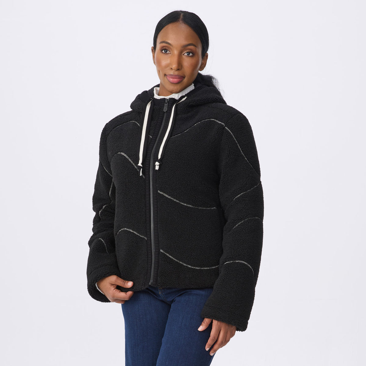A woman modeling a black fleece zip-up hoodie with white drawstrings and contrasting white stitched lines, paired with blue jeans, against a light gray studio background.