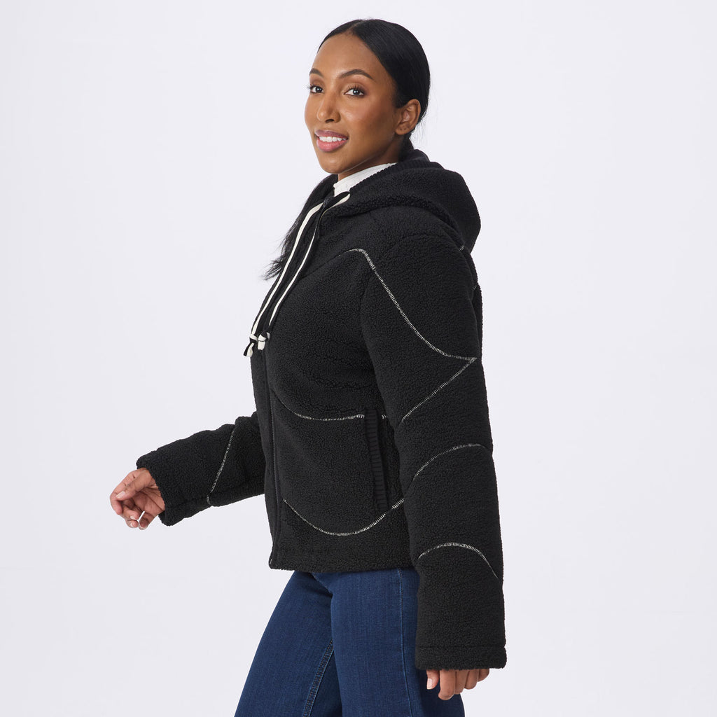 A woman wearing a black  fleece cropped hoodie jacket with white contrast stitching, paired with blue jeans, photographed against a solid light gray background.