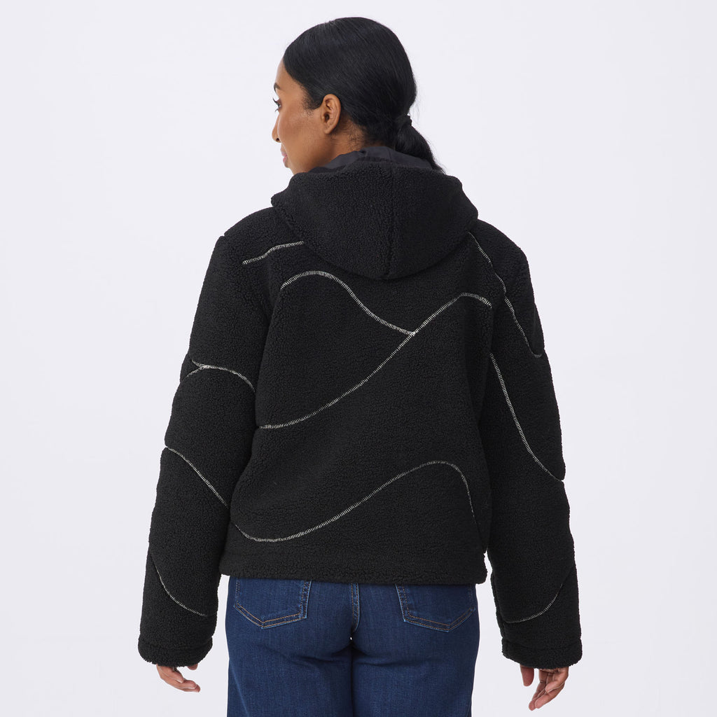 A woman viewed from the back wearing a black fleece hooded jacket with wavy silver contrast stitching, paired with blue jeans, against a plain light gray studio background.