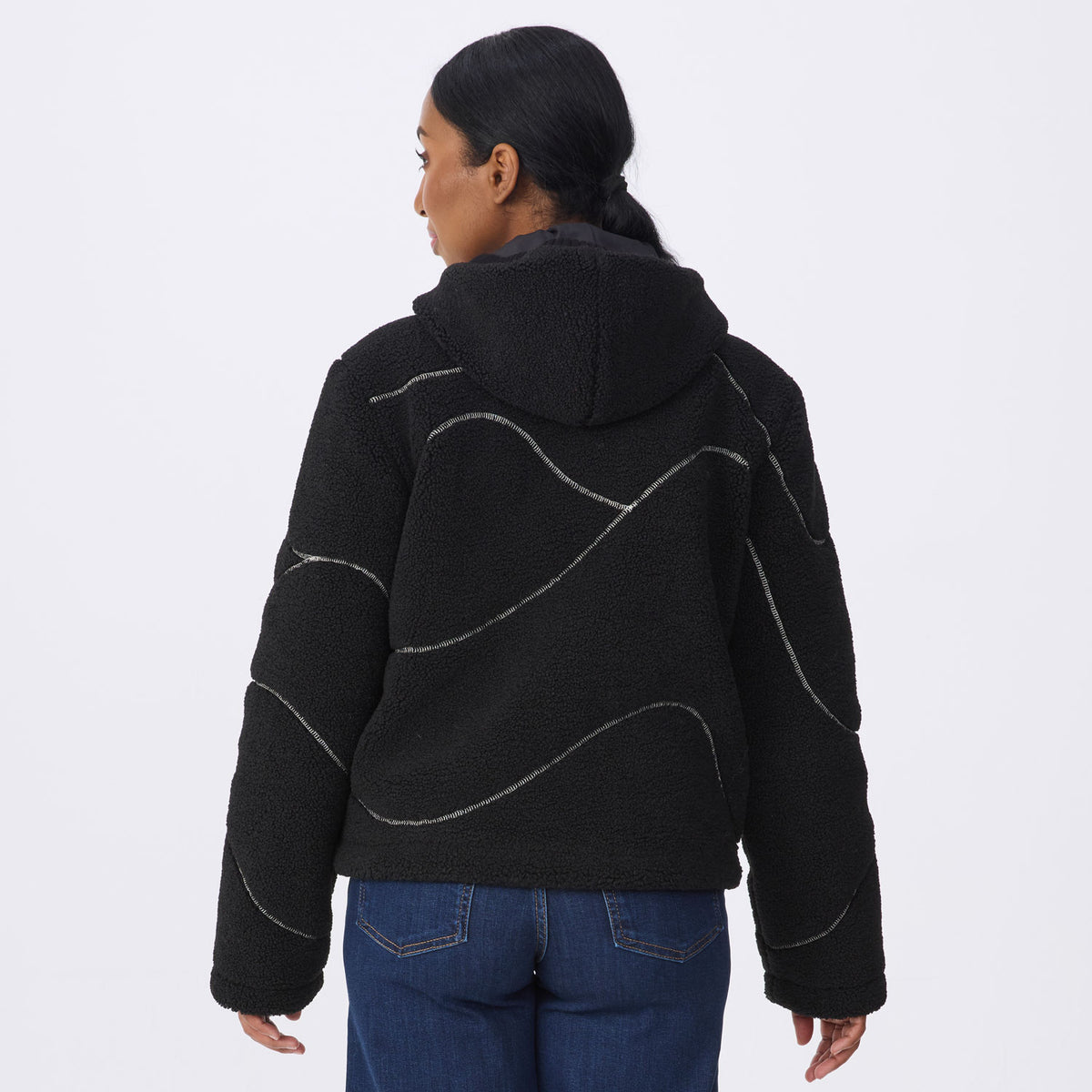 A woman viewed from the back wearing a black fleece hooded jacket with wavy silver contrast stitching, paired with blue jeans, against a plain light gray studio background.