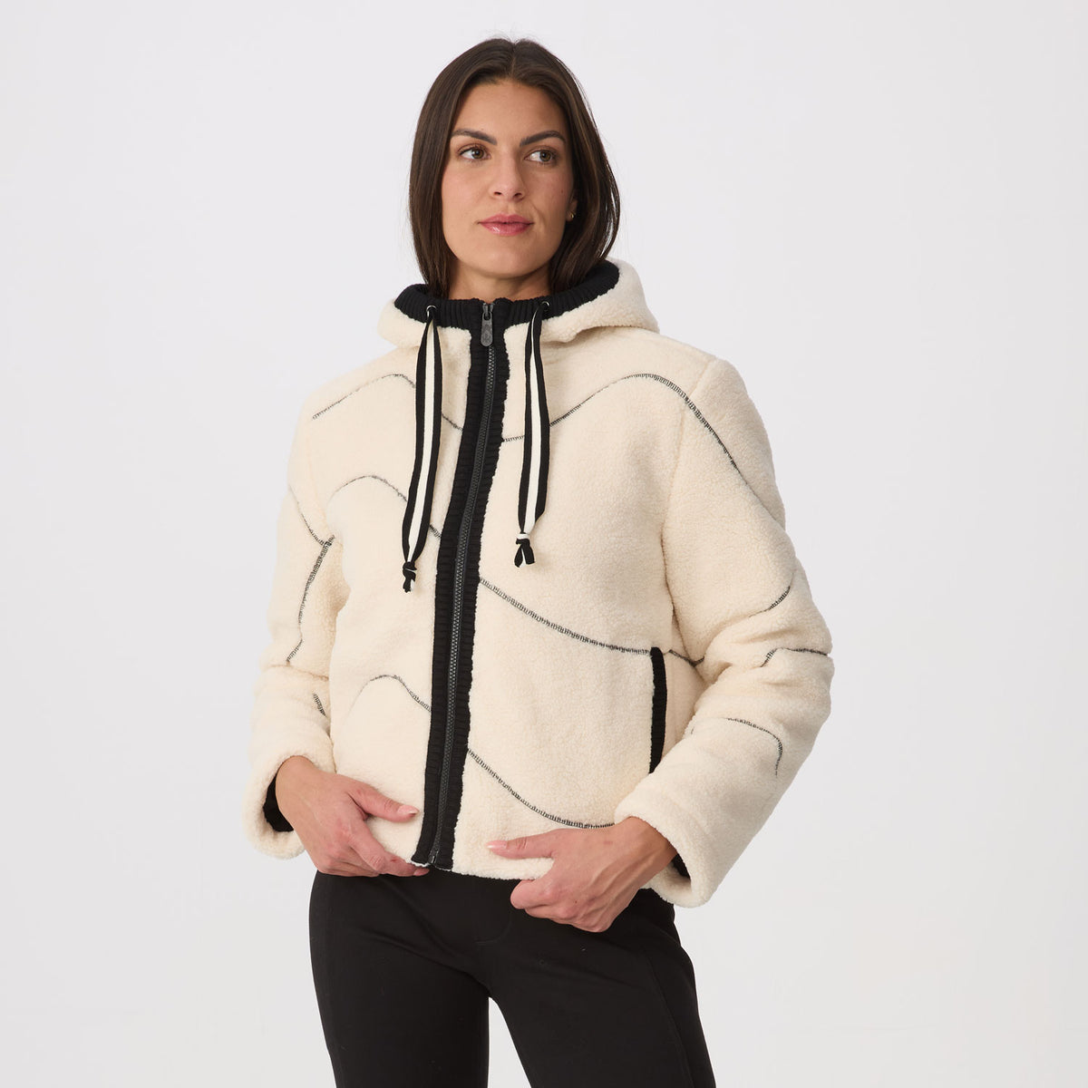 A woman modeling a cropped, fleece hooded jacket in cream with black zippers, drawstrings, and black side detailing, paired with black pants, against a plain light gray studio background.