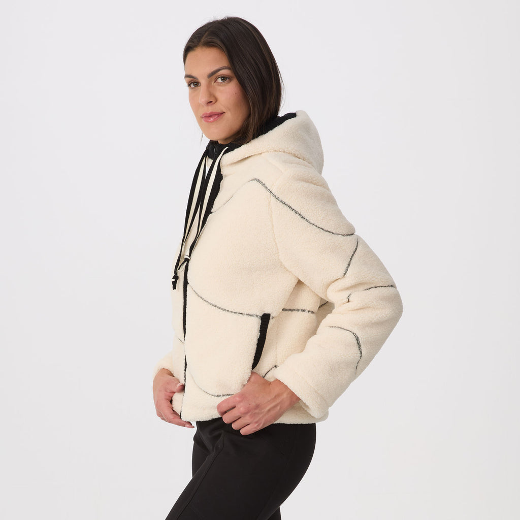 A woman wearing a cream-colored fleece cropped hoodie jacket with black contrast stitching and black drawstrings, paired with black pants, against a plain white studio background.