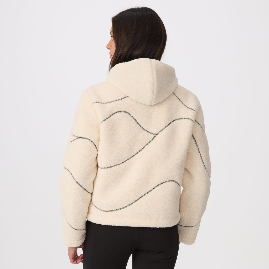 A woman shown from the back wearing a cream-colored fleece cropped hoodie with dark gray wavy line patterns across the back, paired with black pants, against a plain white studio background.