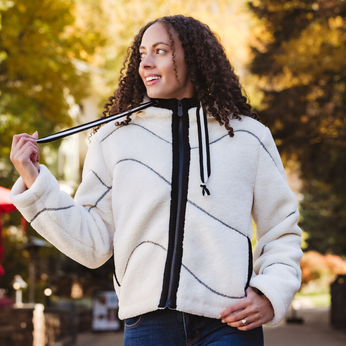 A woman wearing a cream-colored fleece zip-up hoodie jacket with black contrast stitching and a black front zipper, standing outdoors in a park with trees in the background.