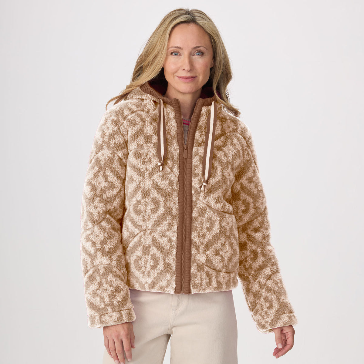 A woman wearing a cropped fleece hooded zip-up jacket with a beige and brown geometric pattern, paired with light beige pants, photographed against a plain light gray studio background.