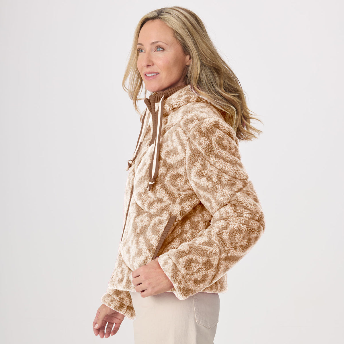 A woman wearing a fleece hooded jacket with a tan and cream leopard-like pattern, shown in profile against a plain light gray studio background.