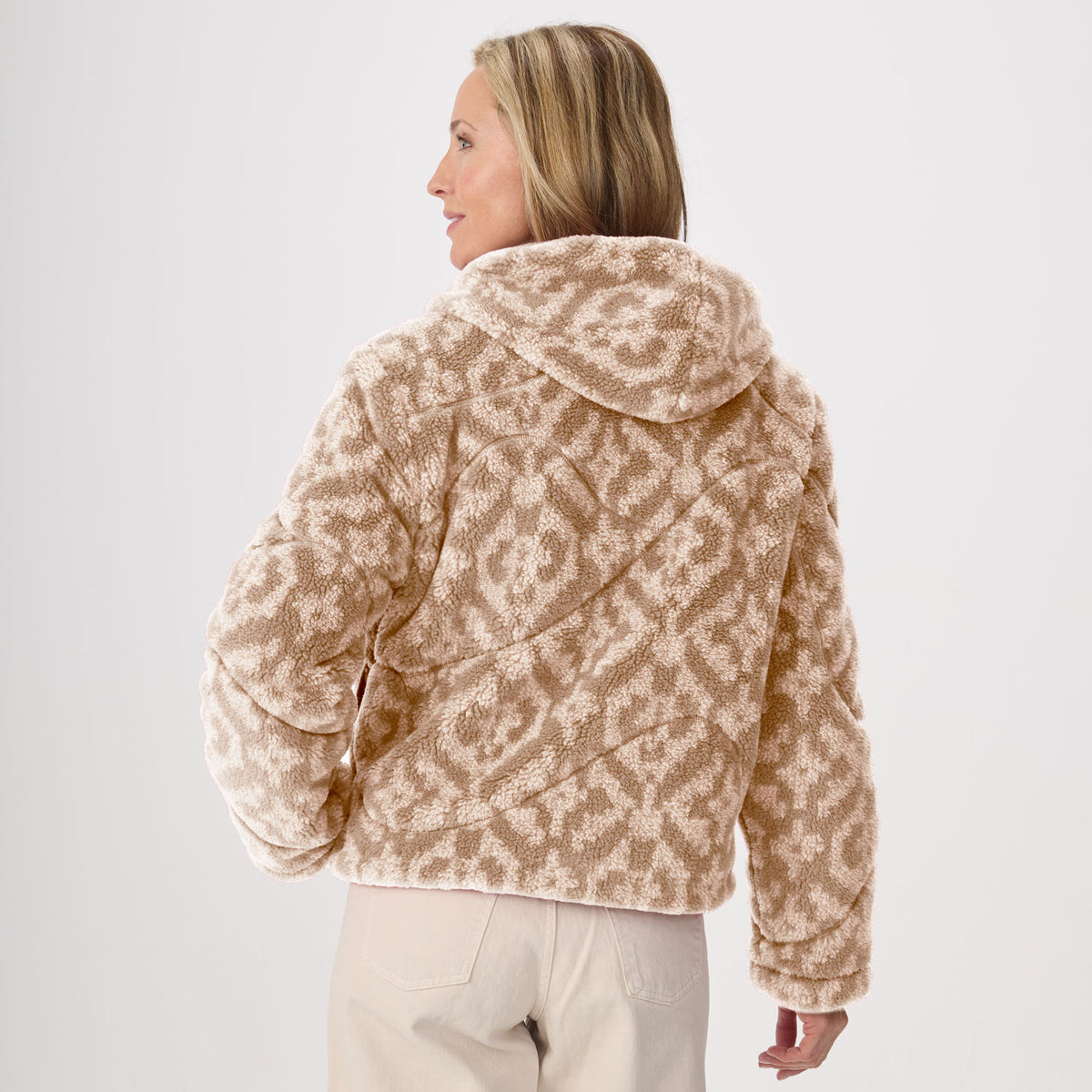 A woman shown from the back wearing a cropped, fleece hooded jacket with a beige and off-white geometric pattern, paired with light beige pants, against a plain light gray studio background.