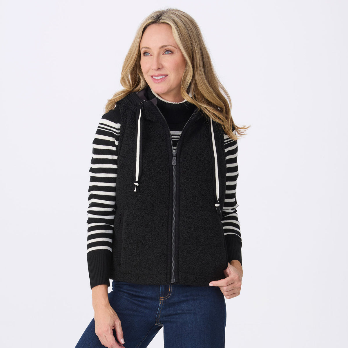 A woman modeling a black zip-up fleece vest with white drawstrings, paired with a black turtleneck and blue jeans, against a light gray studio background.