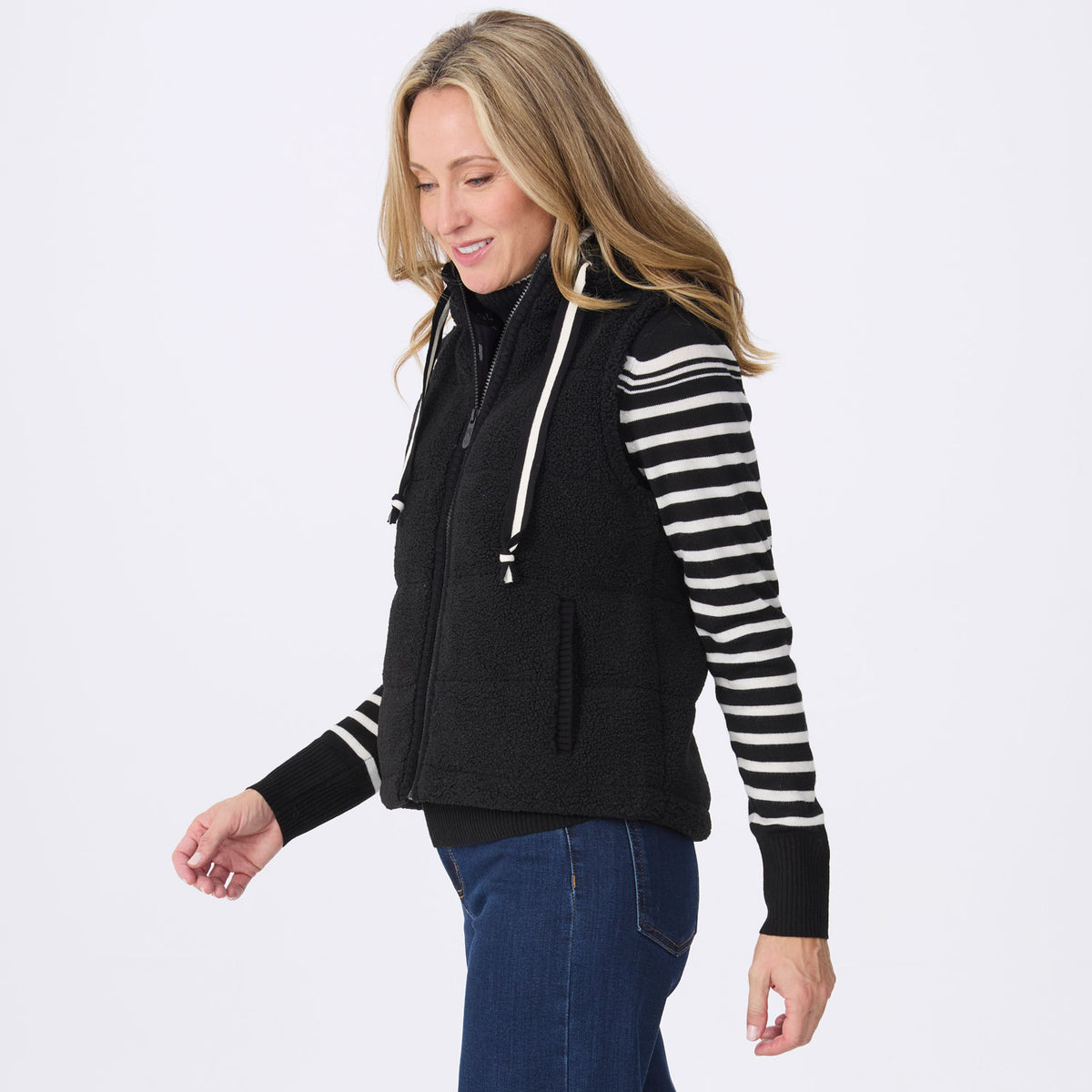 A woman wearing a black fleece zip-front vest with white drawstrings over a black-and-white striped long-sleeve top and blue jeans, photographed against a clean white studio background.