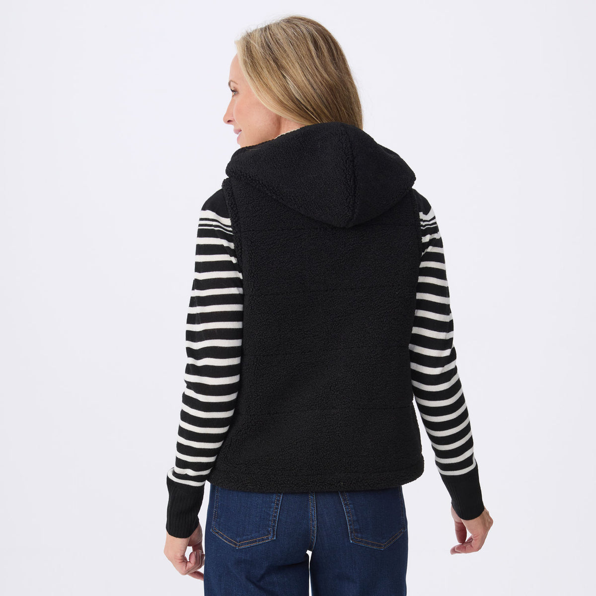A woman shown from the back wearing a black fleece hooded vest over a black and white striped long-sleeve top, paired with blue jeans, against a plain white studio background.