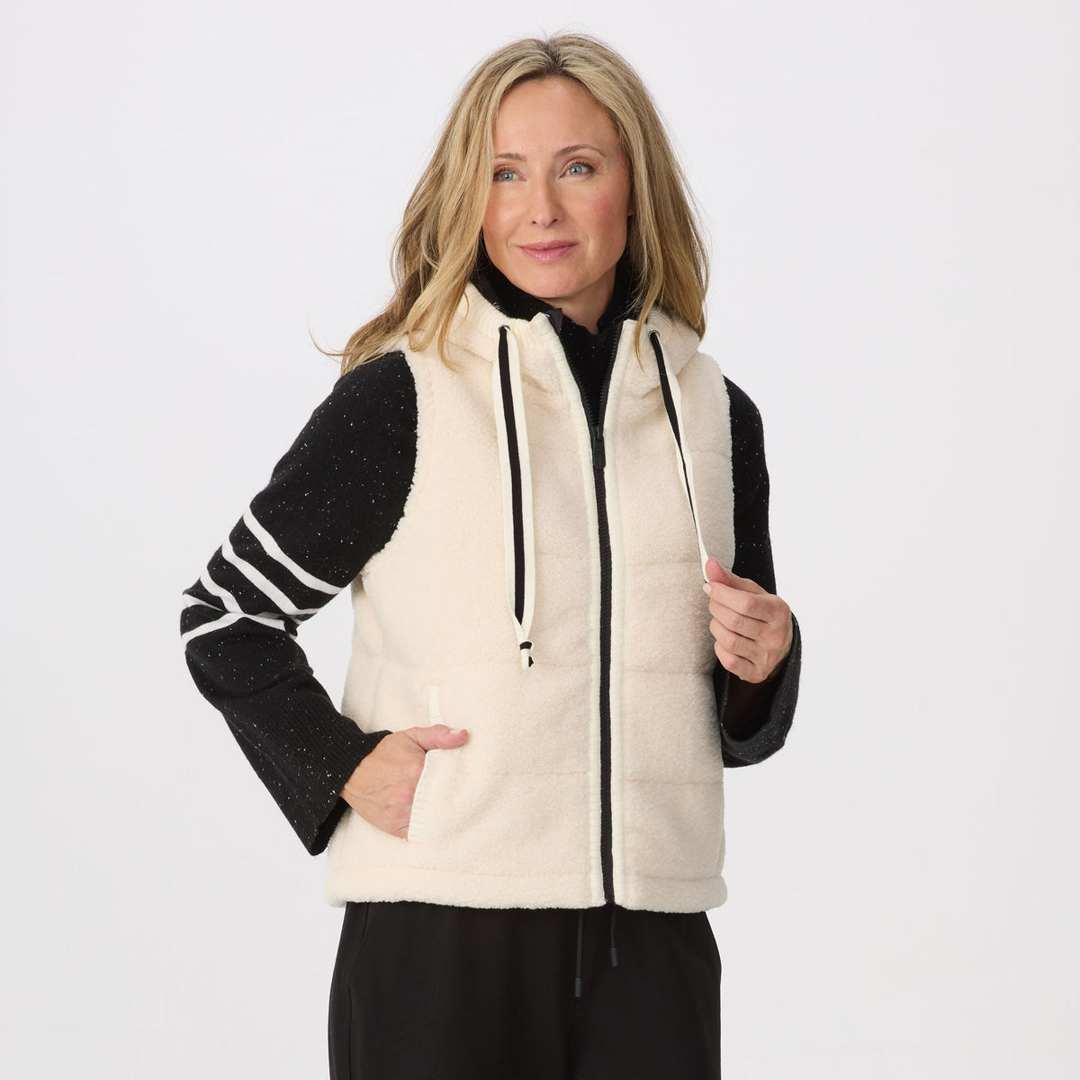 A woman modeling a cream fleece zip-front vest with black trim over a black long-sleeve top with white stripes on the sleeves, against a light gray studio background.