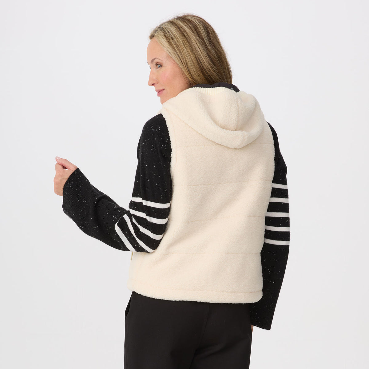 A woman shown from the back wearing a cream fleece hooded vest over a black knit top with white stripes on the sleeves, against a plain light gray studio background.