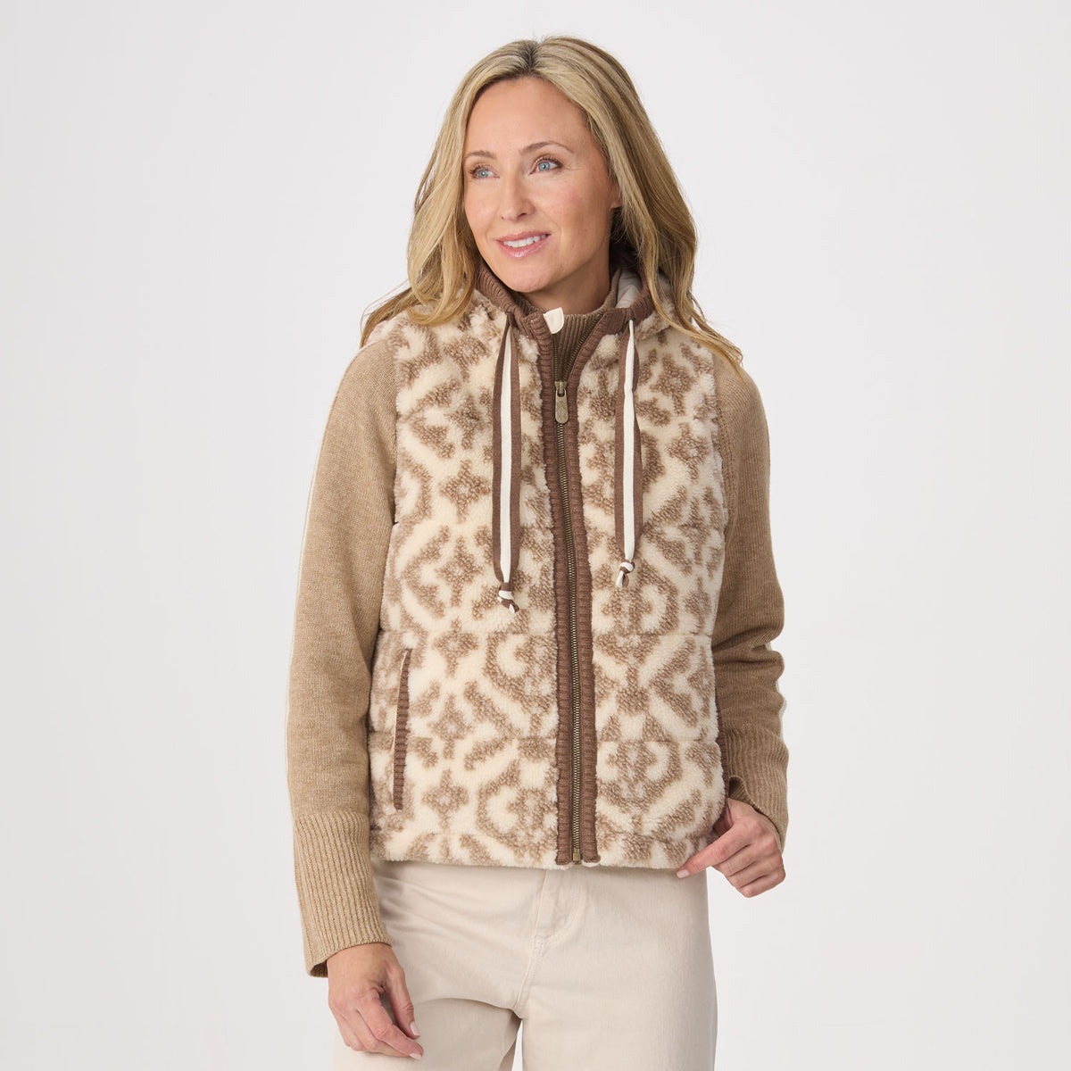 A woman modeling a zip-front fleece vest with a brown and beige geometric pattern, tan ribbed sleeves, and a brown zipper, paired with light beige pants against a plain light gray studio background.