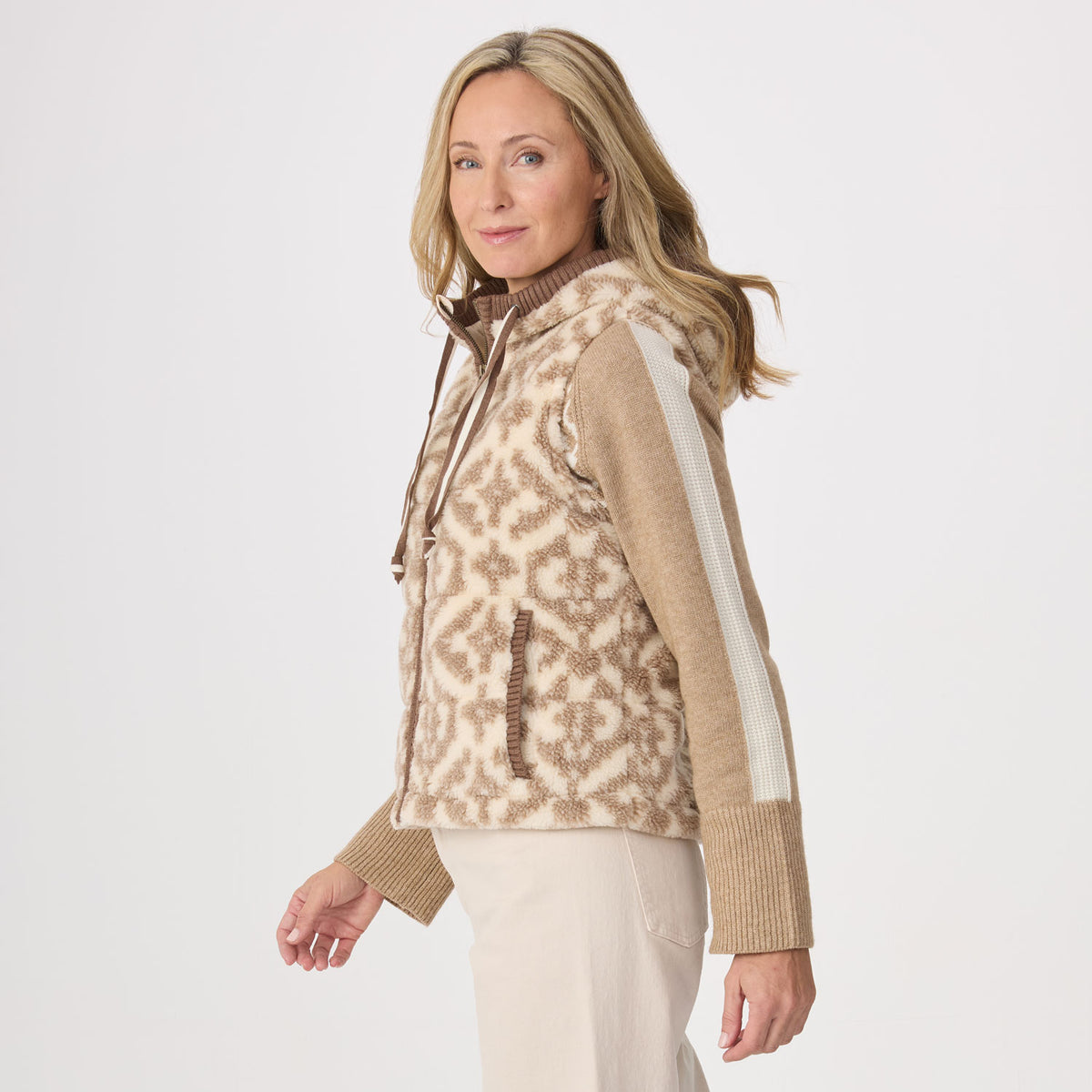 A woman modeling a fleece sleeveless zip-front vest with a beige and brown abstract pattern, ribbed brown trim, and light beige sleeves, photographed against a clean light gray studio background.