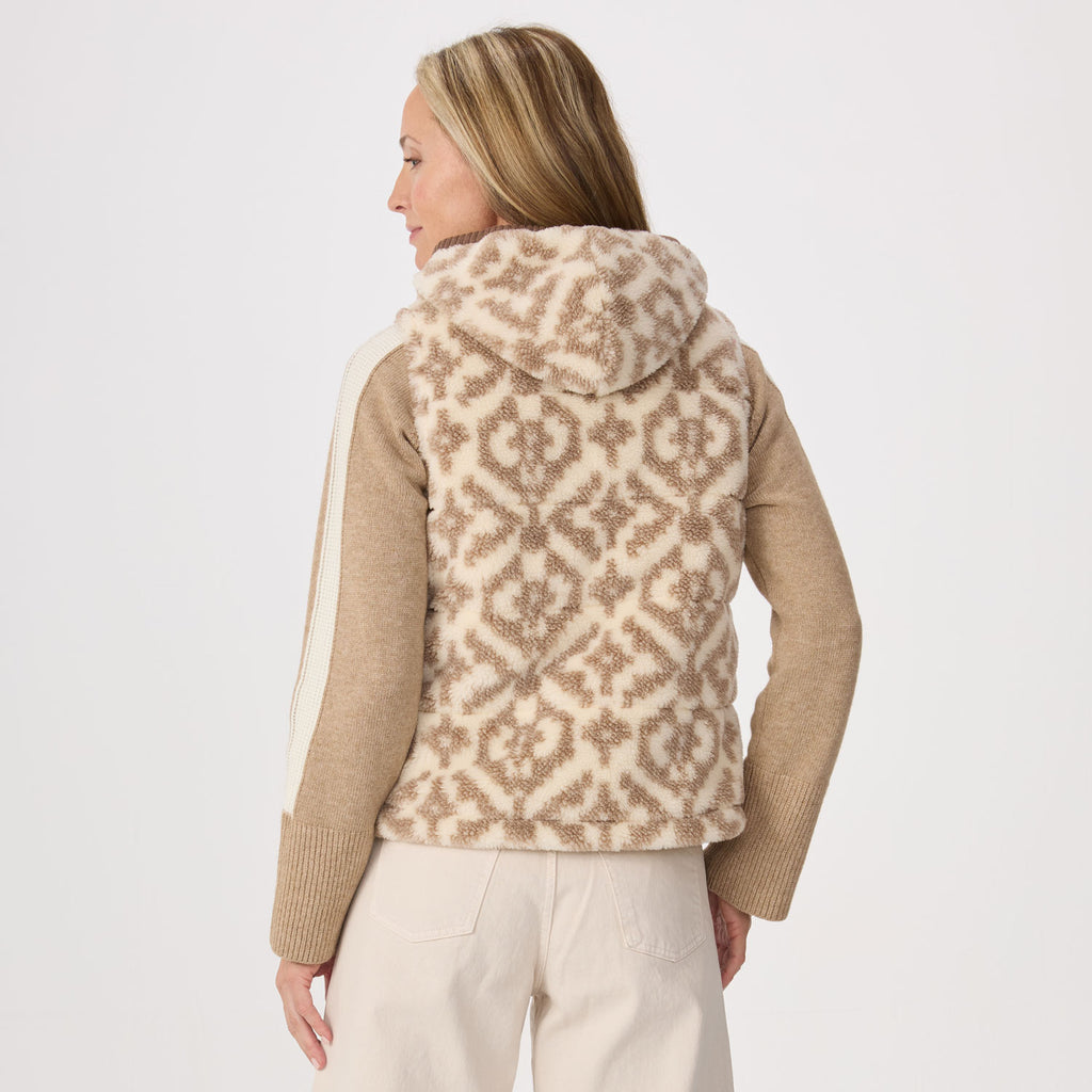A woman shown from the back wearing a fleece beige and cream patterned hooded vest over a tan sweater and cream pants, against a plain light gray studio background.