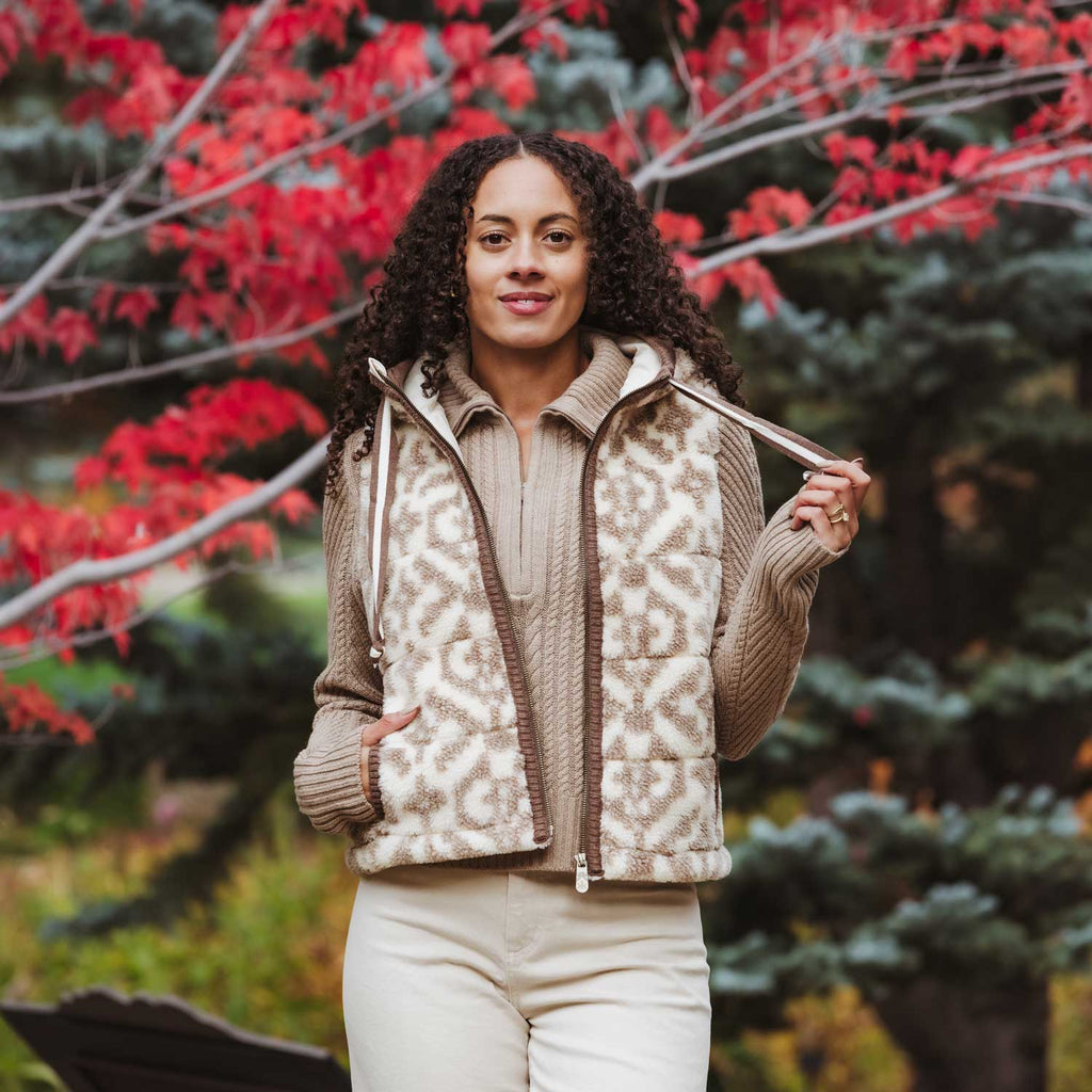 A woman wearing a fleece beige and brown geometric patterned zip-up vest over a matching knit quarter-zip sweater and cream pants, standing outdoors in front of a tree with bright red autumn leaves.