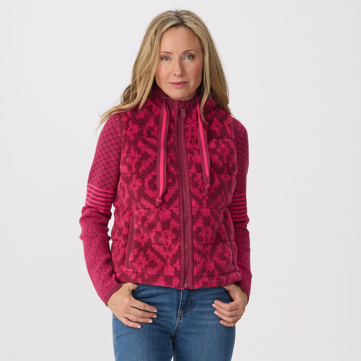 A woman modeling a bright pink and magenta patterned fleece knit zip-up vest with blue jeans, photographed against a solid light gray background.