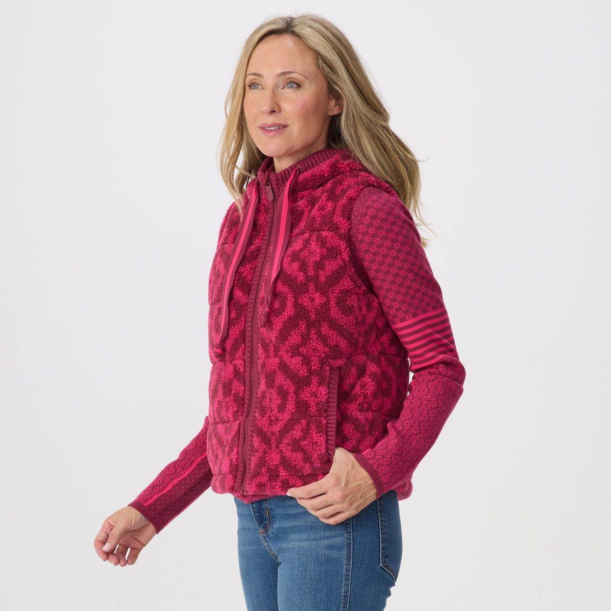 A woman modeling a pink and magenta patterned fleece zip-up hoodie vest, paired with blue jeans, against a light gray studio background.