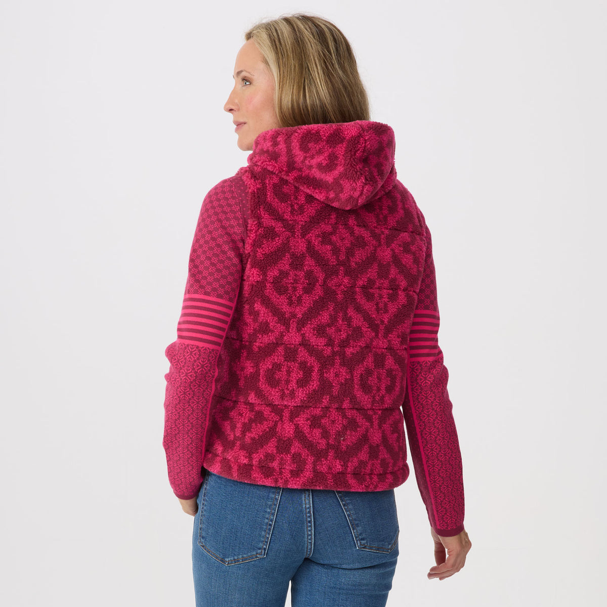 A woman shown from the back wearing a bright pink hooded vest with a darker pink geometric pattern, paired with blue jeans against a light gray studio background.