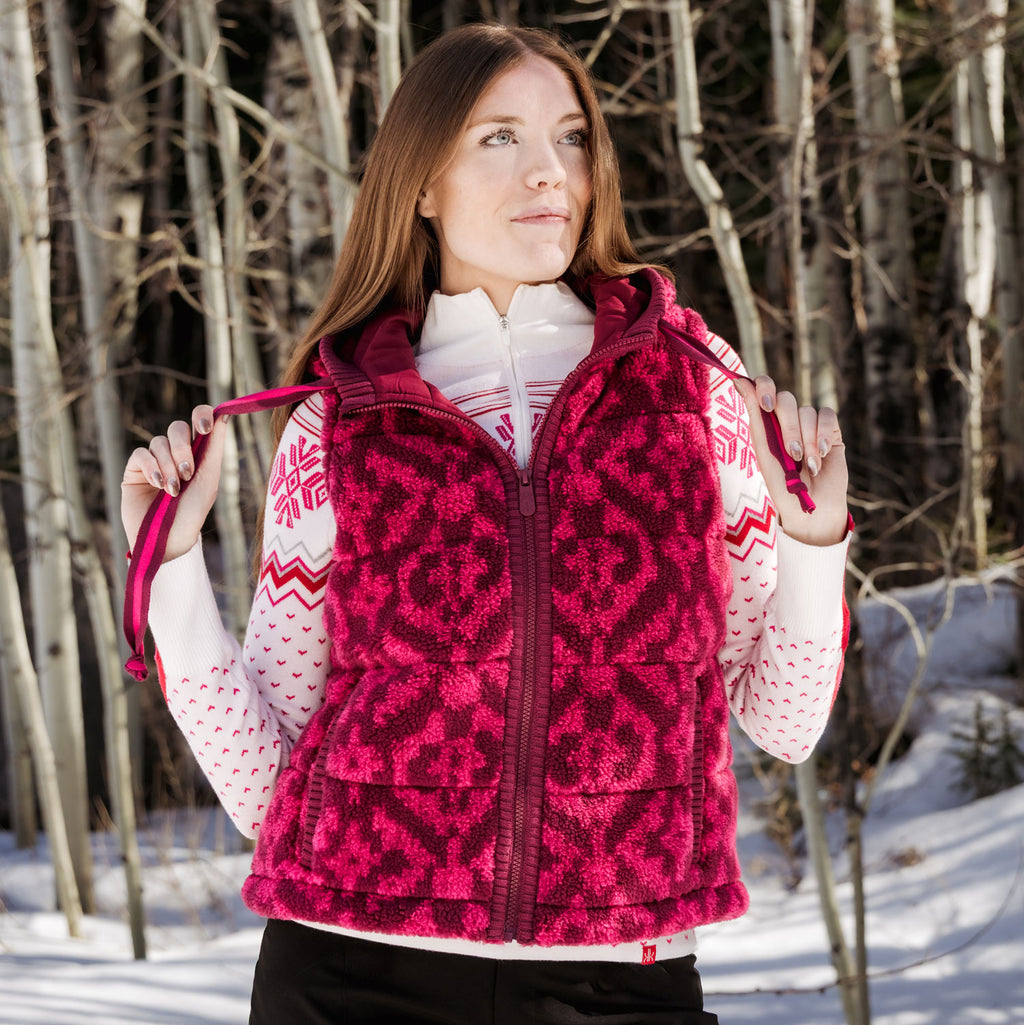 A woman wearing a bright pink and dark red patterned fleece vest over a white patterned sweater, standing in a snowy outdoor setting with bare trees in the background.