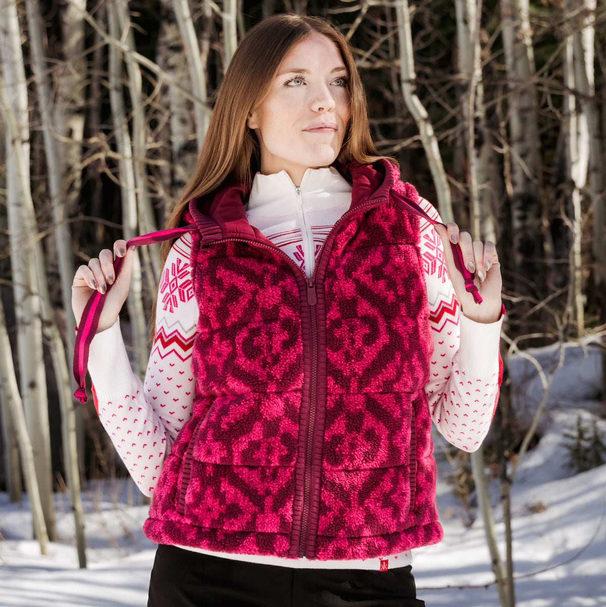 A woman wearing a bright pink and dark red patterned fleece vest over a white patterned sweater, standing in a snowy outdoor setting with bare trees in the background.