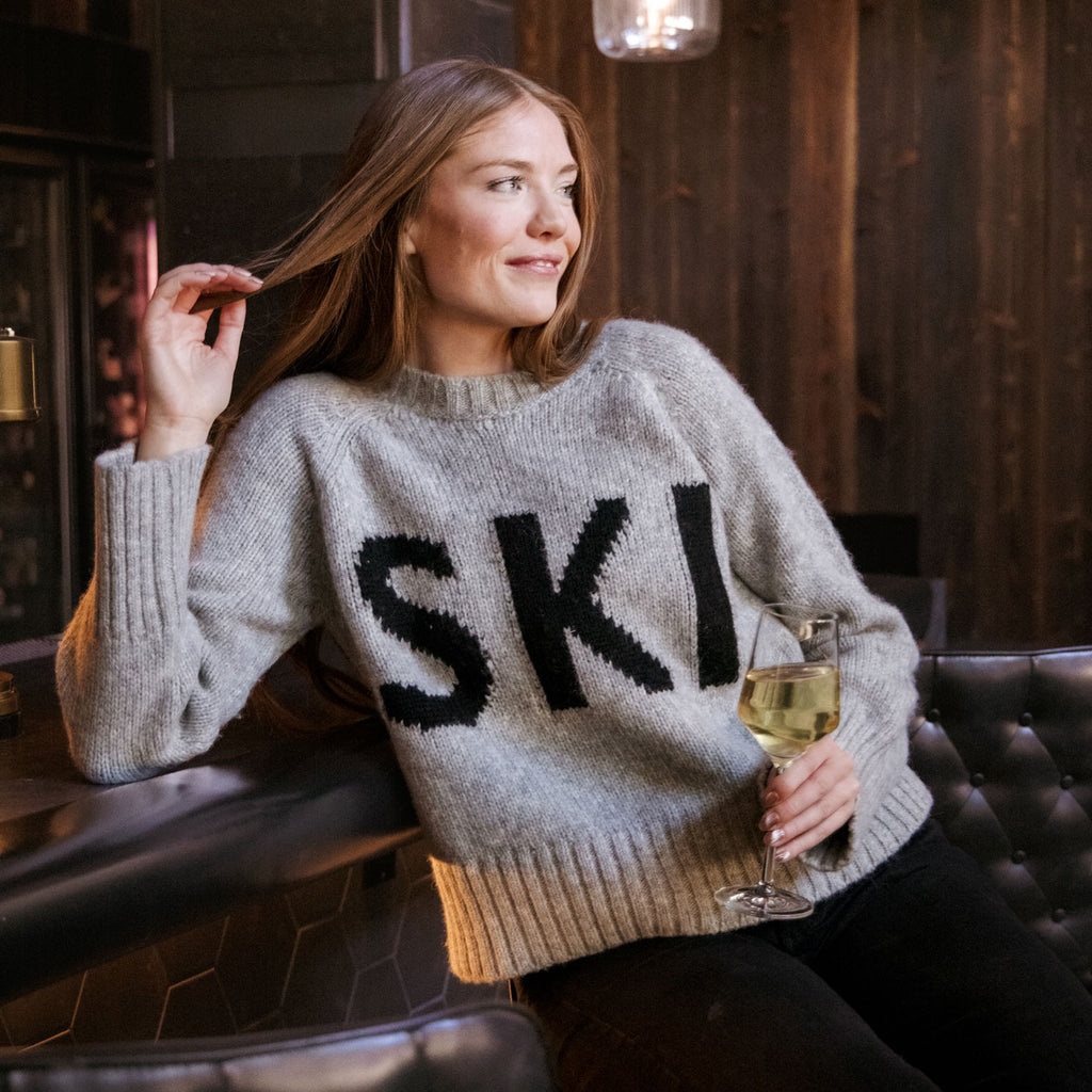 A woman sitting in a dimly lit bar or lounge wearing a light gray knit sweater with large black 'SKI' lettering across the front, holding a glass of white wine.