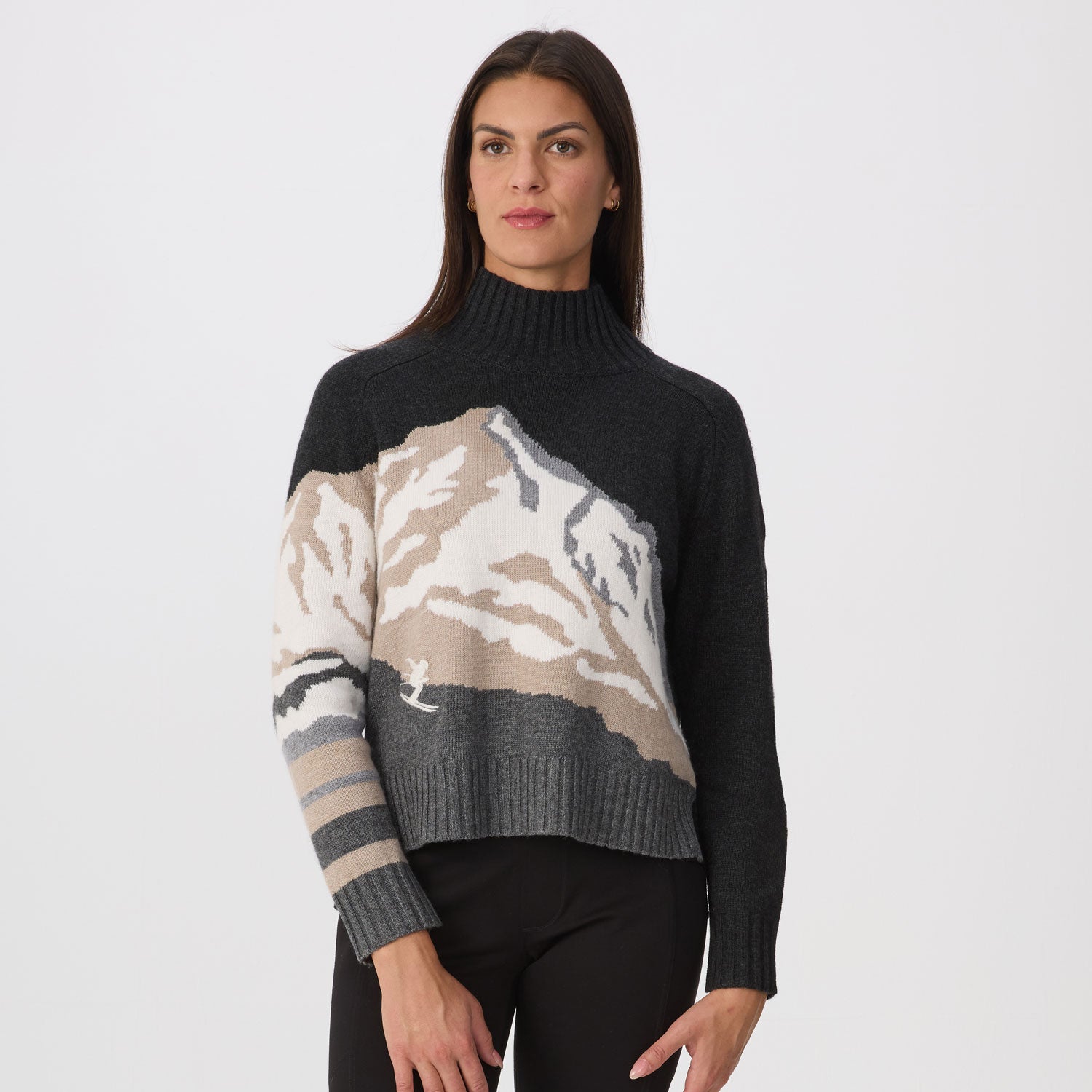 A woman modeling a cropped mock-neck knit sweater with a dark charcoal base and a large mountain graphic across the chest and sleeves in beige, gray, and brown tones, paired with black pants against a light gray studio background.