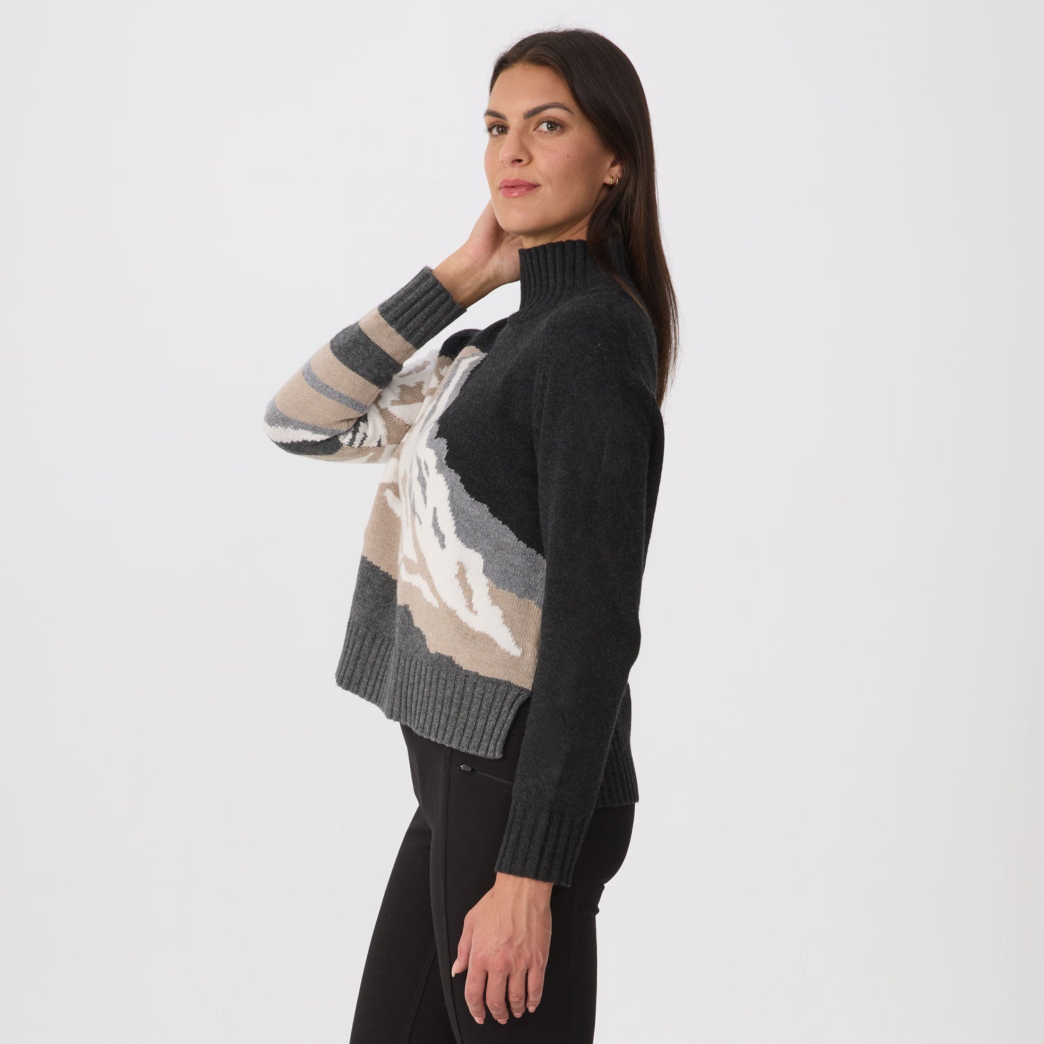 A woman modeling a dark charcoal knit turtleneck sweater with a large abstract mountain pattern in beige, gray, and white across the front, paired with black pants, against a plain light gray studio background.