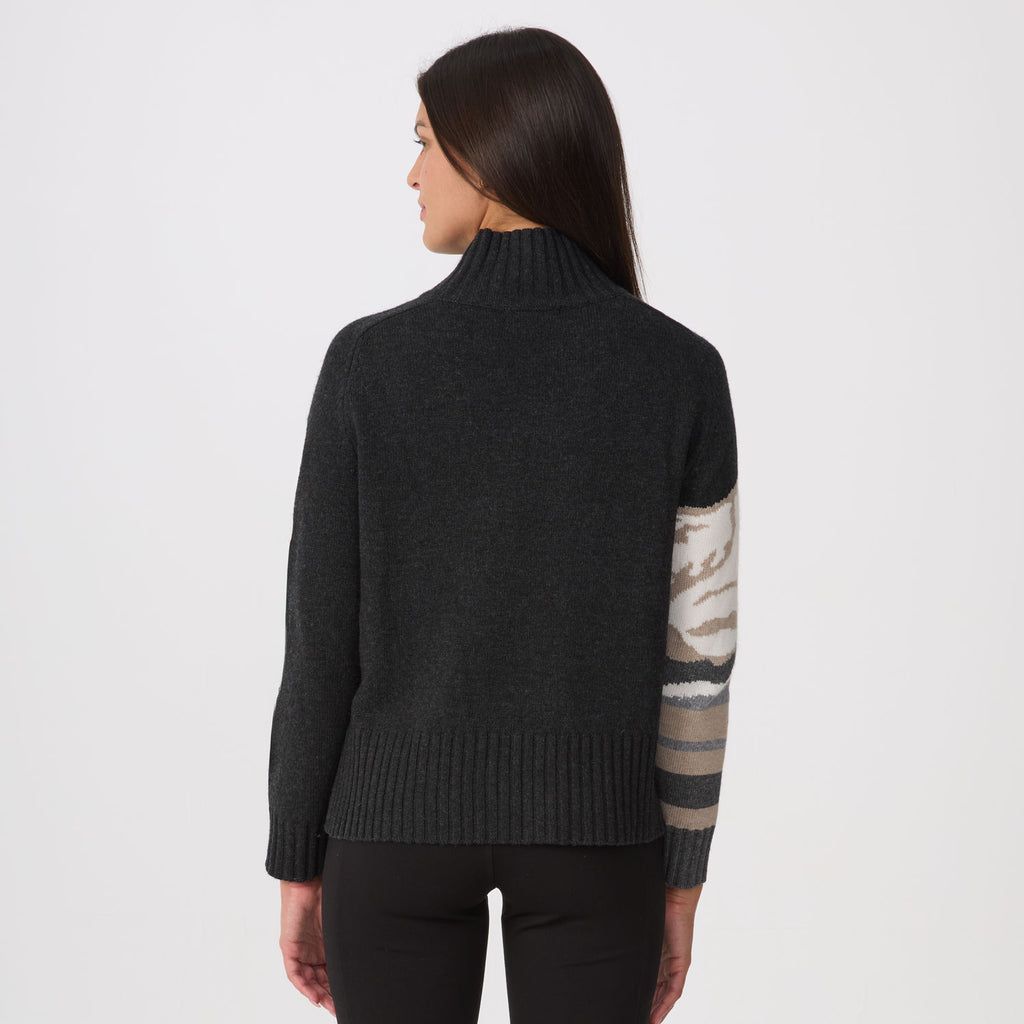 A woman shown from the back wearing a dark charcoal gray ribbed turtleneck sweater with a contrasting beige and gray patterned sleeve, paired with black pants, against a plain white studio background.