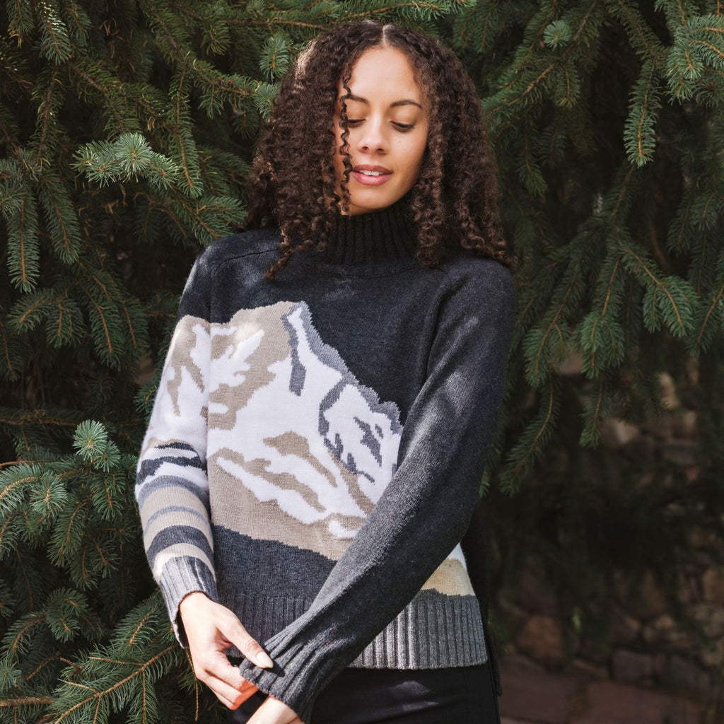 A woman with curly brown hair wearing a dark gray mock-neck knit sweater with a large beige and gray mountain pattern on the front, standing outdoors in front of green pine trees.