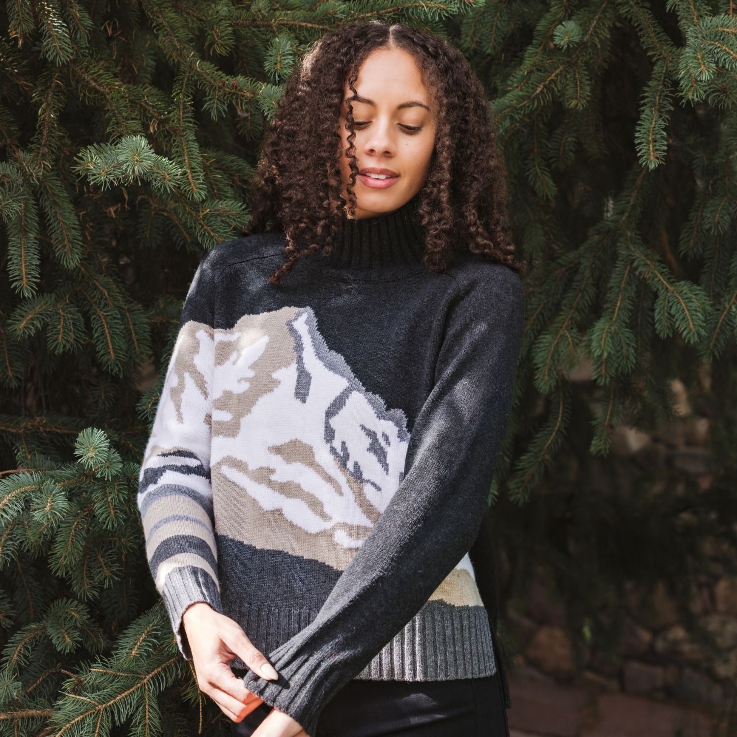 A woman with curly brown hair wearing a dark gray mock-neck knit sweater with a large beige and gray mountain pattern on the front, standing outdoors in front of green pine trees.