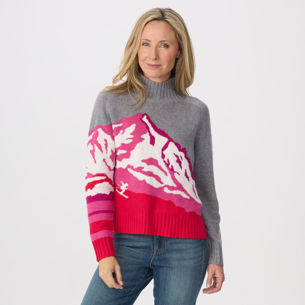 A woman modeling a gray knit turtleneck sweater with a large mountain design in pink, magenta, and white across the chest and one sleeve, paired with blue jeans against a light gray studio background.