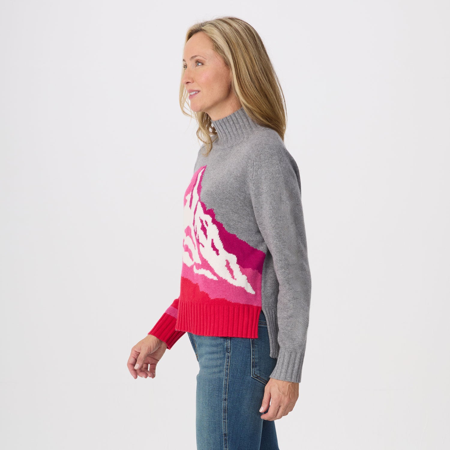 A woman in side profile wearing a gray knit mock-neck sweater with a vibrant pink and white mountain graphic across the front, paired with blue jeans, against a plain white studio background.