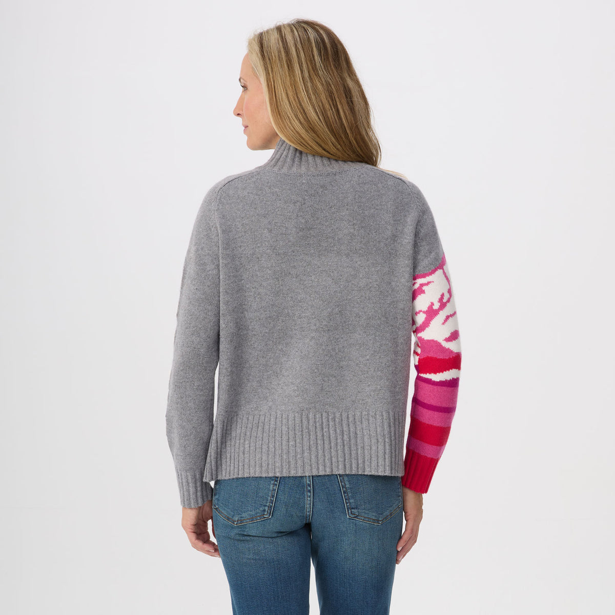 A woman shown from the back wearing a gray knit sweater with a vibrant pink, red, and white patterned sleeve, paired with blue jeans against a plain white background.