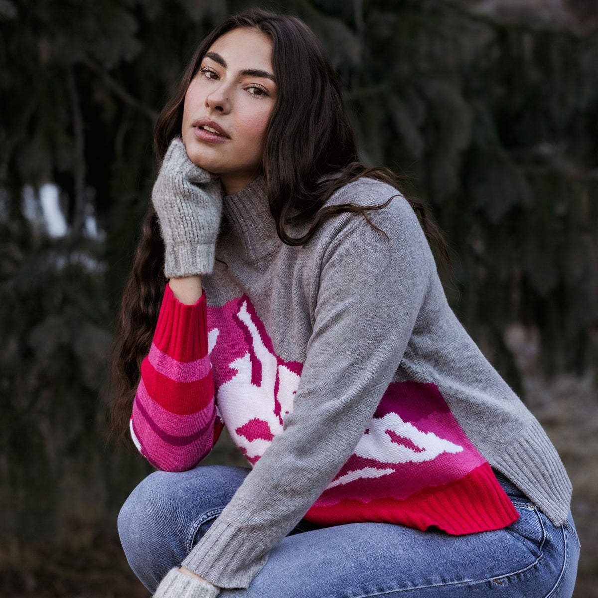A woman wearing a gray knit turtleneck sweater with a pink and white patterned yoke, paired with blue jeans and gray knit mittens, posing outdoors in front of a dark, blurred background of trees.