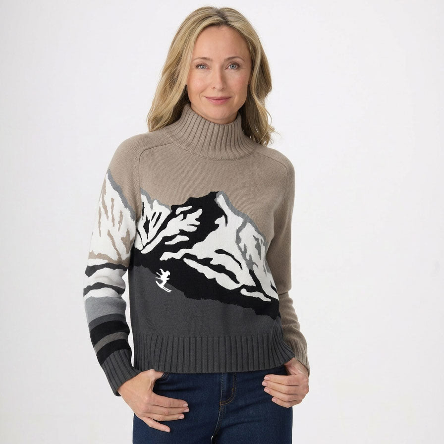 A woman wearing a beige turtleneck sweater with a large black, white, and gray mountain graphic across the chest and sleeves, paired with dark blue jeans, against a plain white studio background.