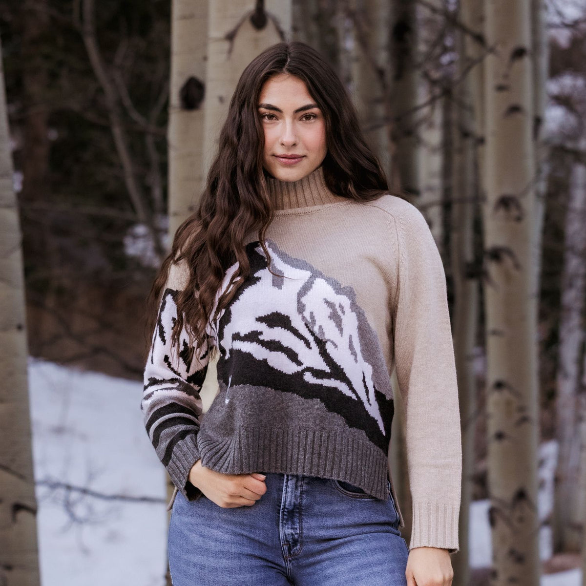 A woman wearing a beige knit turtleneck sweater with a mountain pattern in black, white, and gray, paired with blue jeans, standing in a snowy forest of birch trees.