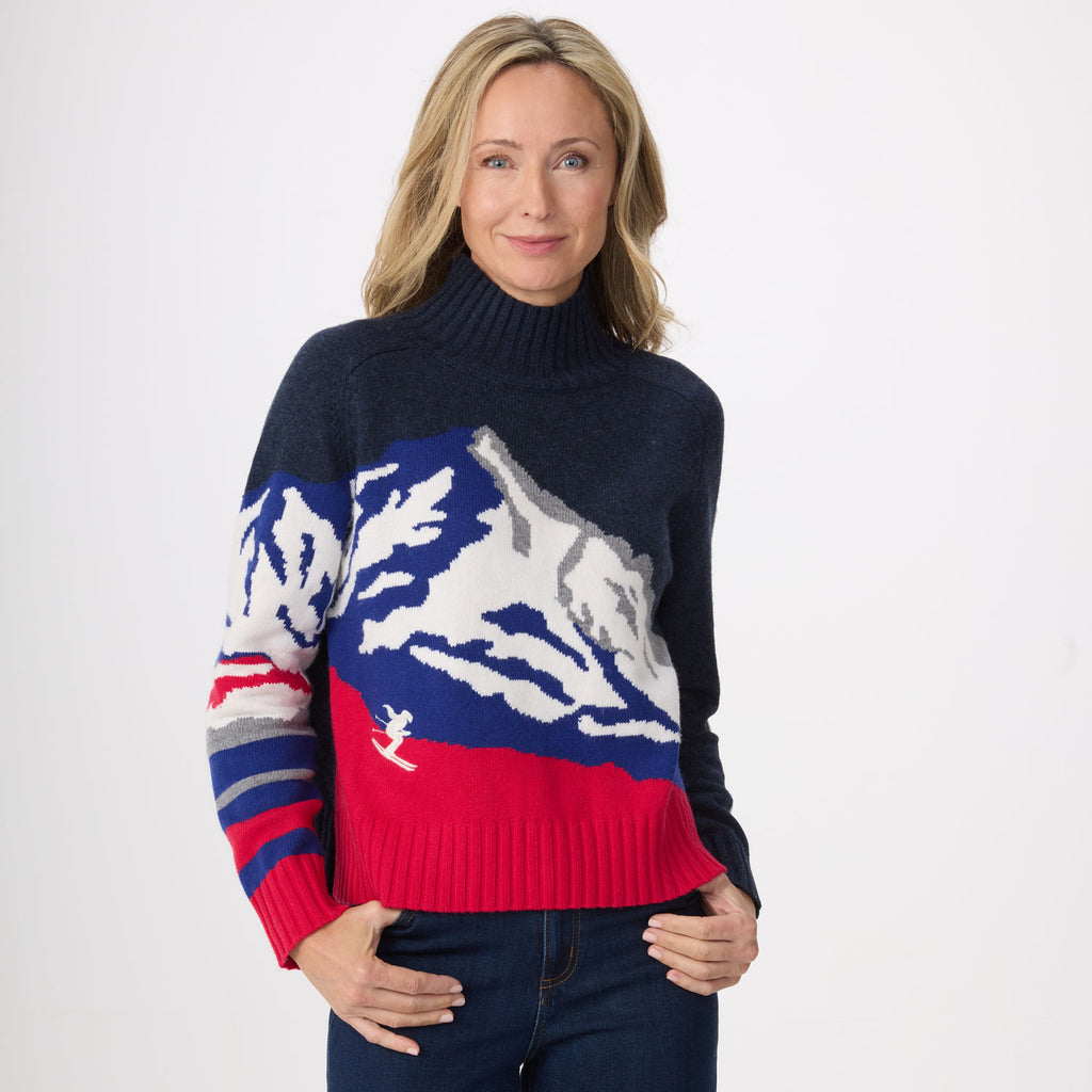 A woman modeling a navy blue knit turtleneck sweater with a large mountain graphic in white, blue, and red across the chest and sleeves, paired with dark blue jeans against a plain white studio background.
