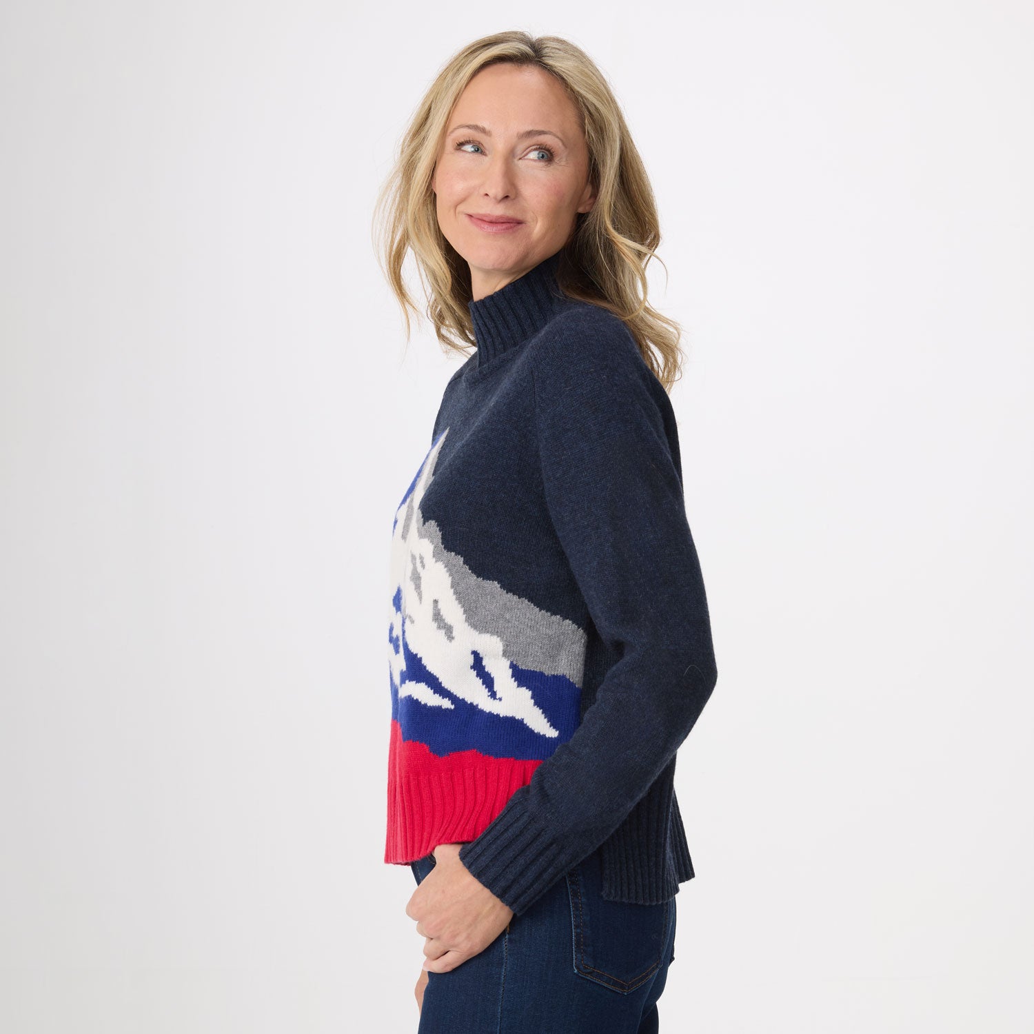 A woman wearing a navy blue knit sweater with a colorful mountain pattern in white, gray, blue, and red, paired with dark blue jeans, photographed against a plain white studio background.