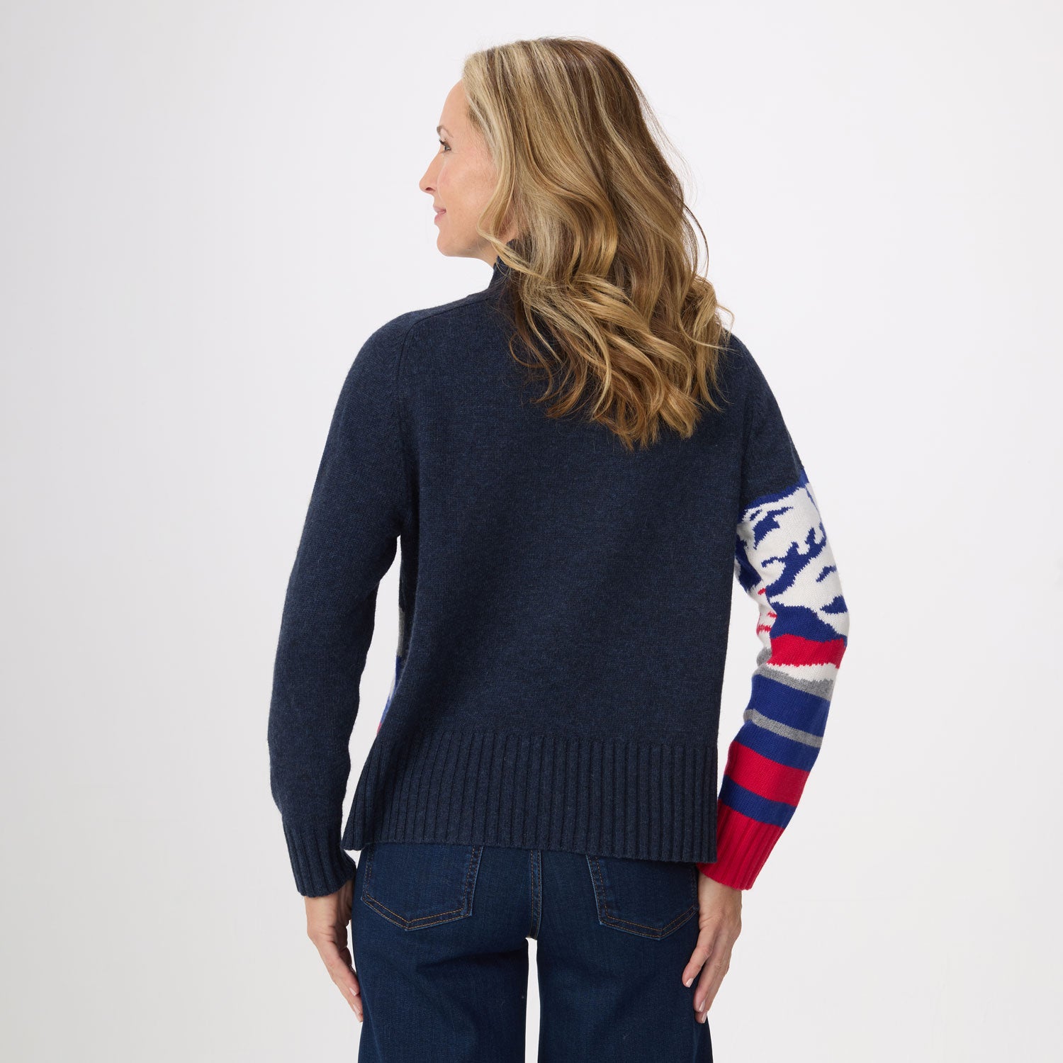 A woman shown from the back wearing a navy blue knit sweater with a multicolor patterned sleeve featuring blue, white, red, and gray stripes, paired with dark blue jeans against a light gray studio background.