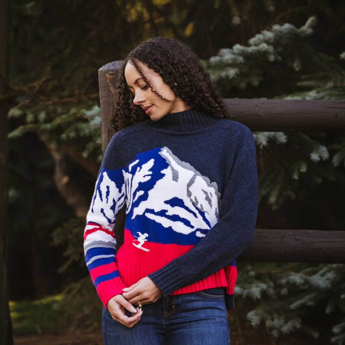 A woman wearing a navy blue knit sweater with a large white mountain graphic and red and blue striped sleeves, standing outdoors in front of a wooden fence and evergreen trees.