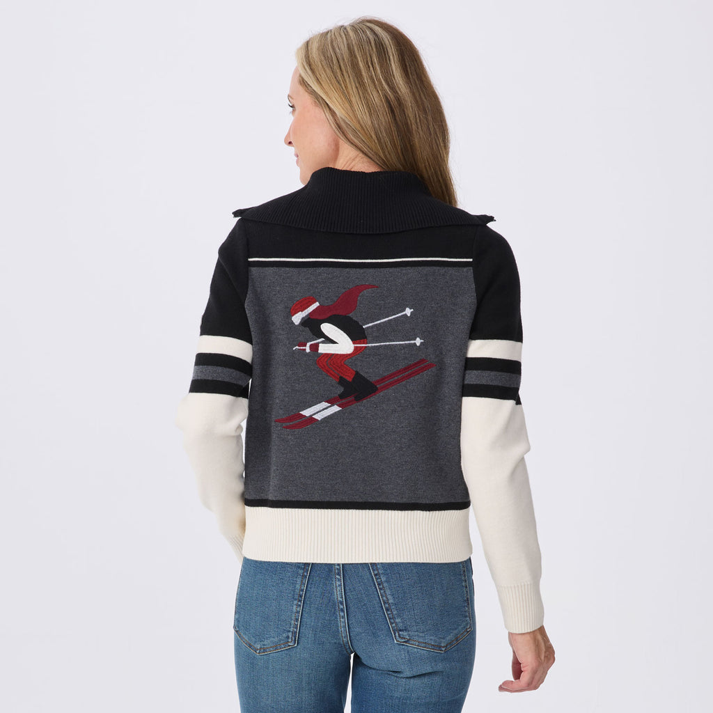 A woman viewed from the back wearing a color-blocked knit sweater with a dark navy collar and shoulders, a dark gray center panel, and off-white sleeves and hem, paired with blue jeans against a light gray studio background.