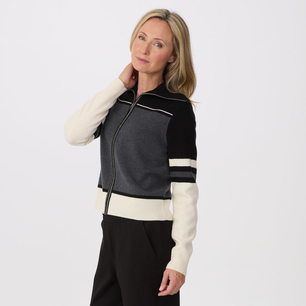 A woman modeling a color-blocked knit zip-up jacket with dark gray, black, and cream panels, paired with black pants, against a plain light gray studio background.