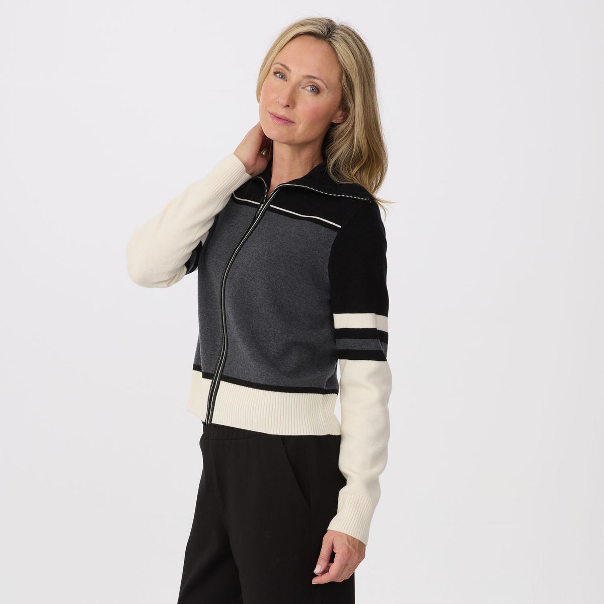 A woman modeling a color-blocked knit zip-up jacket with dark gray, black, and cream panels, paired with black pants, against a plain light gray studio background.