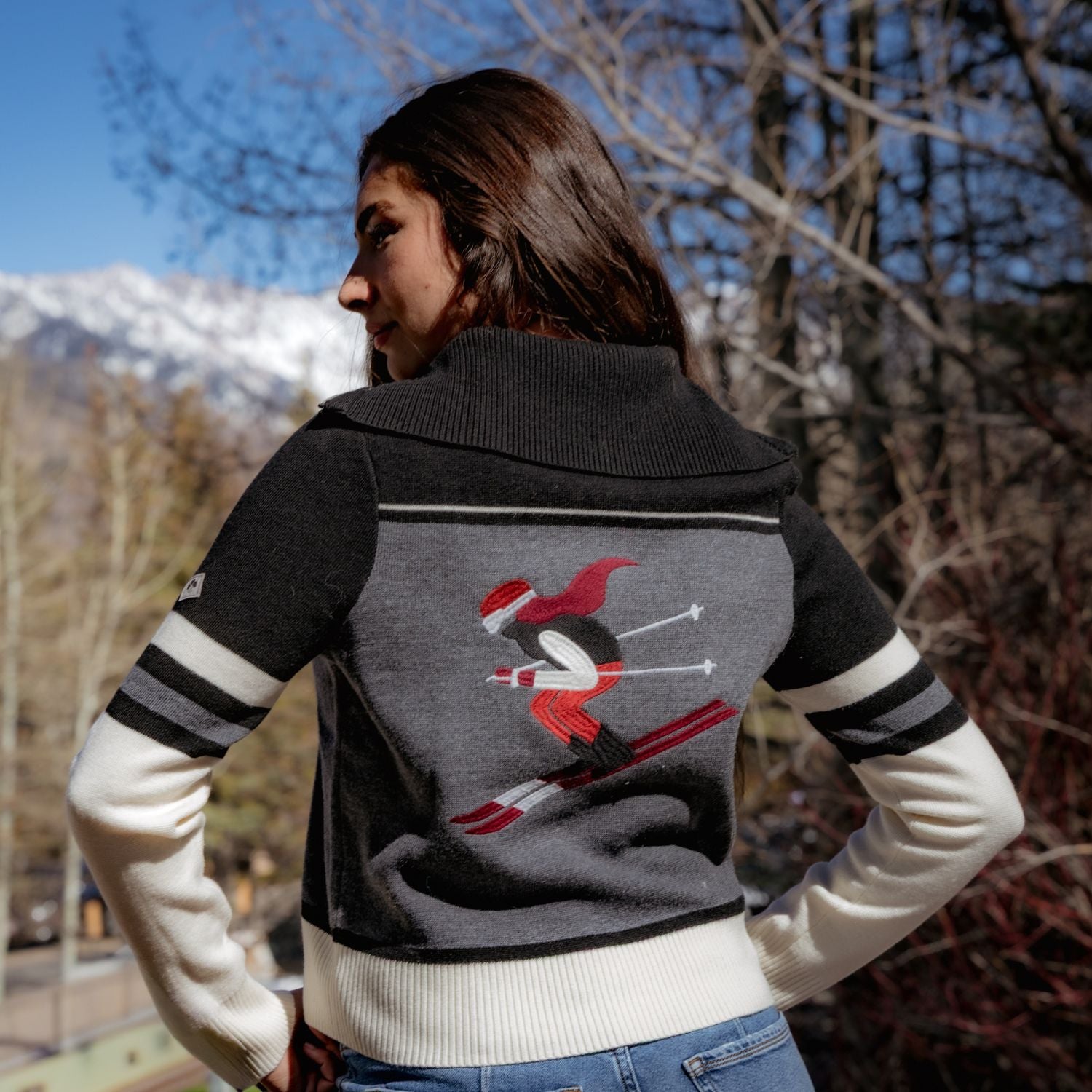 A woman wearing a dark gray and white knit sweater with a red and black skier graphic on the back, photographed outdoors with mountains and trees in the background.