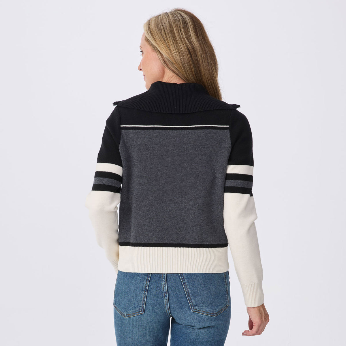 A woman viewed from the back wearing a color-blocked knit sweater with a dark navy collar and shoulders, a dark gray center panel, and off-white sleeves and hem, paired with blue jeans against a light gray studio background.
