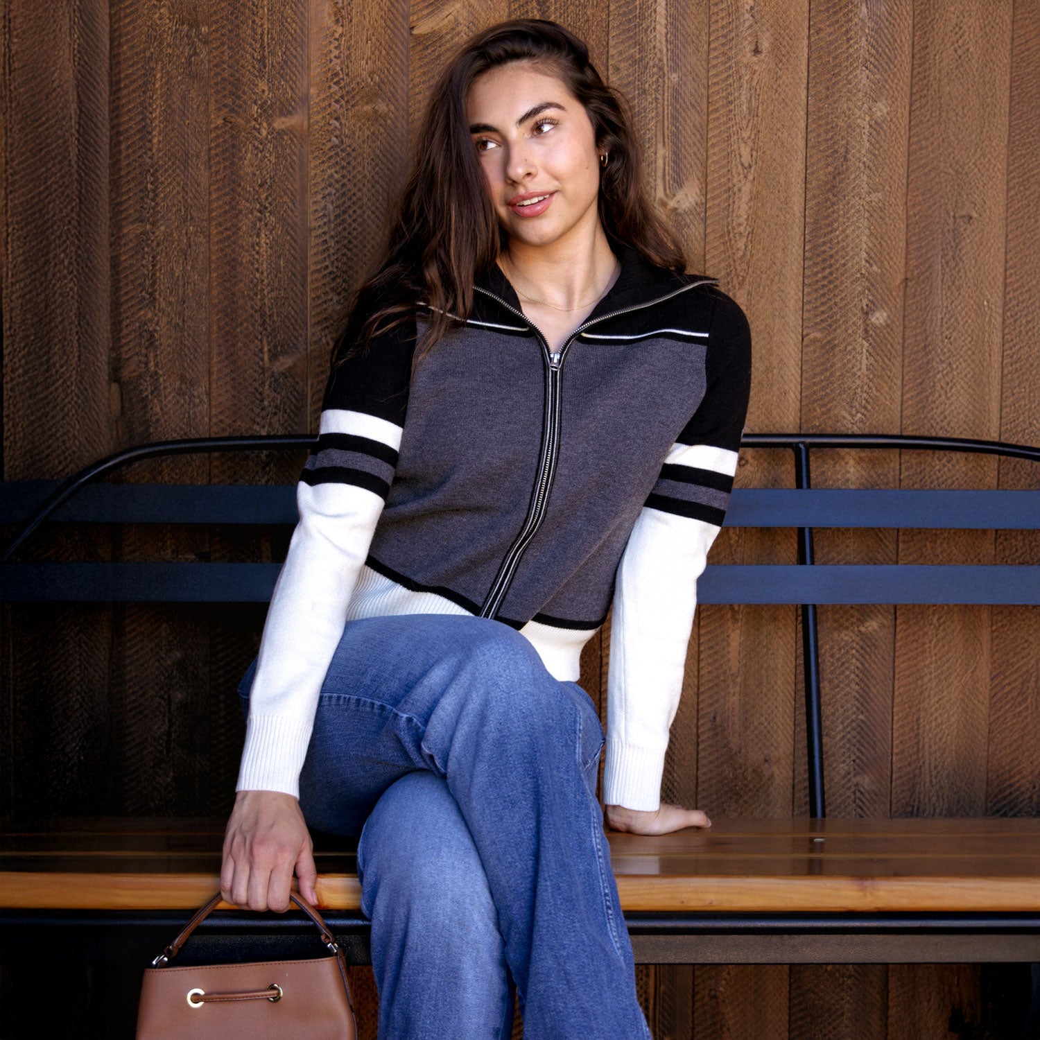 A woman sits on a wooden bench wearing a dark gray and black color-blocked zip-up sweater with white sleeve accents, paired with blue jeans and holding a brown handbag.