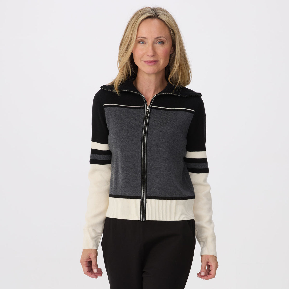 A woman modeling a cropped zip-up knit sweater with a color-blocked design featuring black, dark gray, and cream sections, paired with black pants against a light gray studio background.