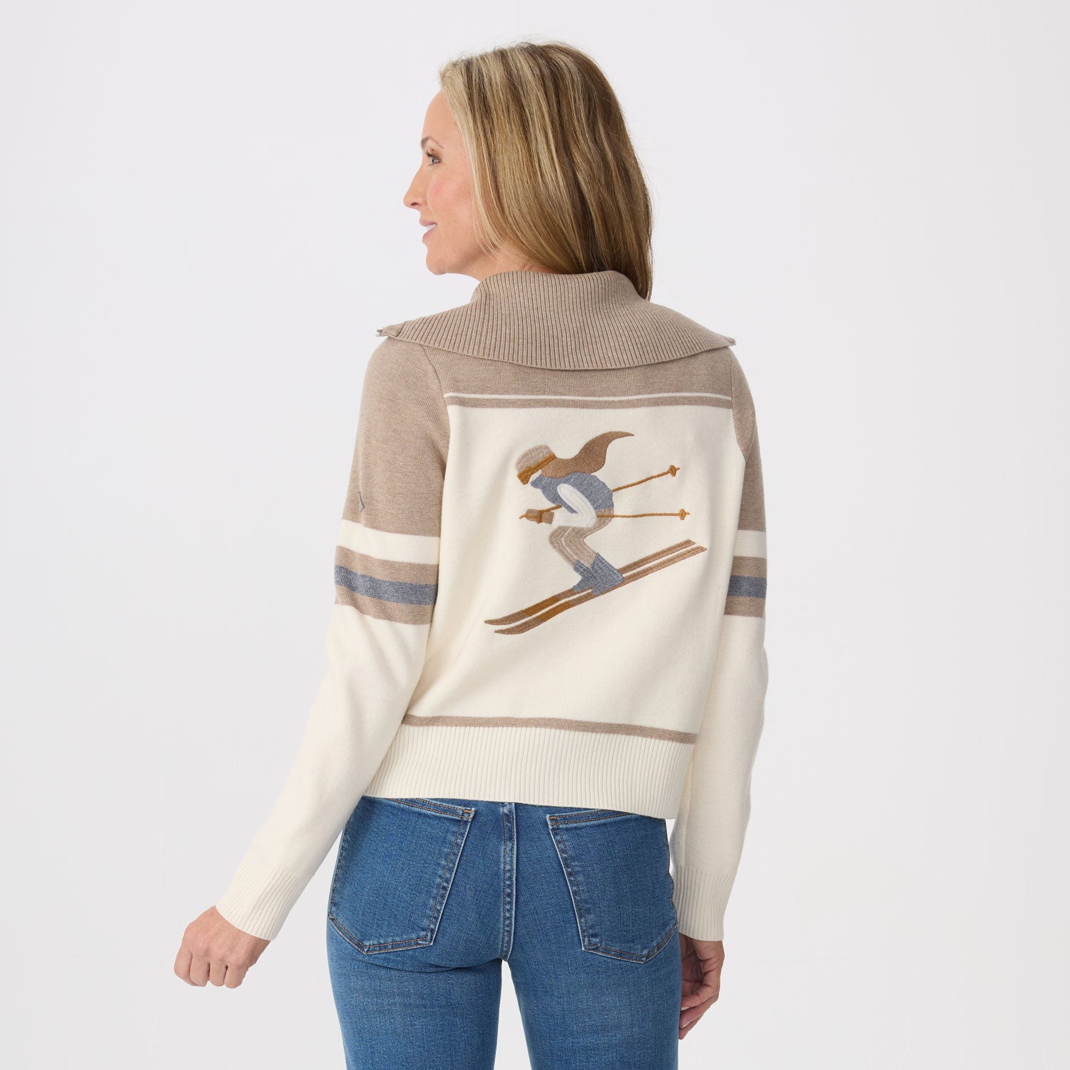 A woman shown from the back wearing a cream and taupe knit sweater with a skier graphic on the back, paired with blue jeans, against a light gray studio background.